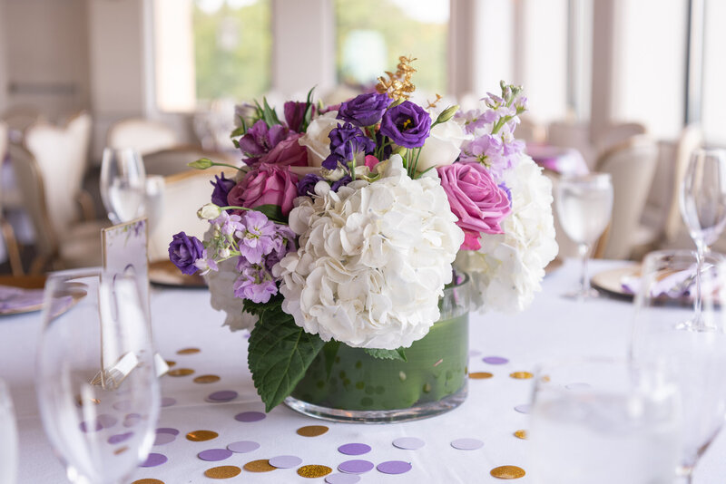 The floral centerpiece on a table with white, purple, and pink flowers at a reception celebration event.