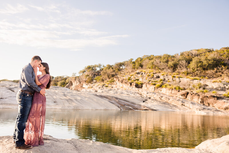 Couple embrace in front of the water during their engagement session at Pedernales Falls State Park in Johnson City, Texas. Photo taken by Austin Engagement Photographers, Joanna & Brett Photography