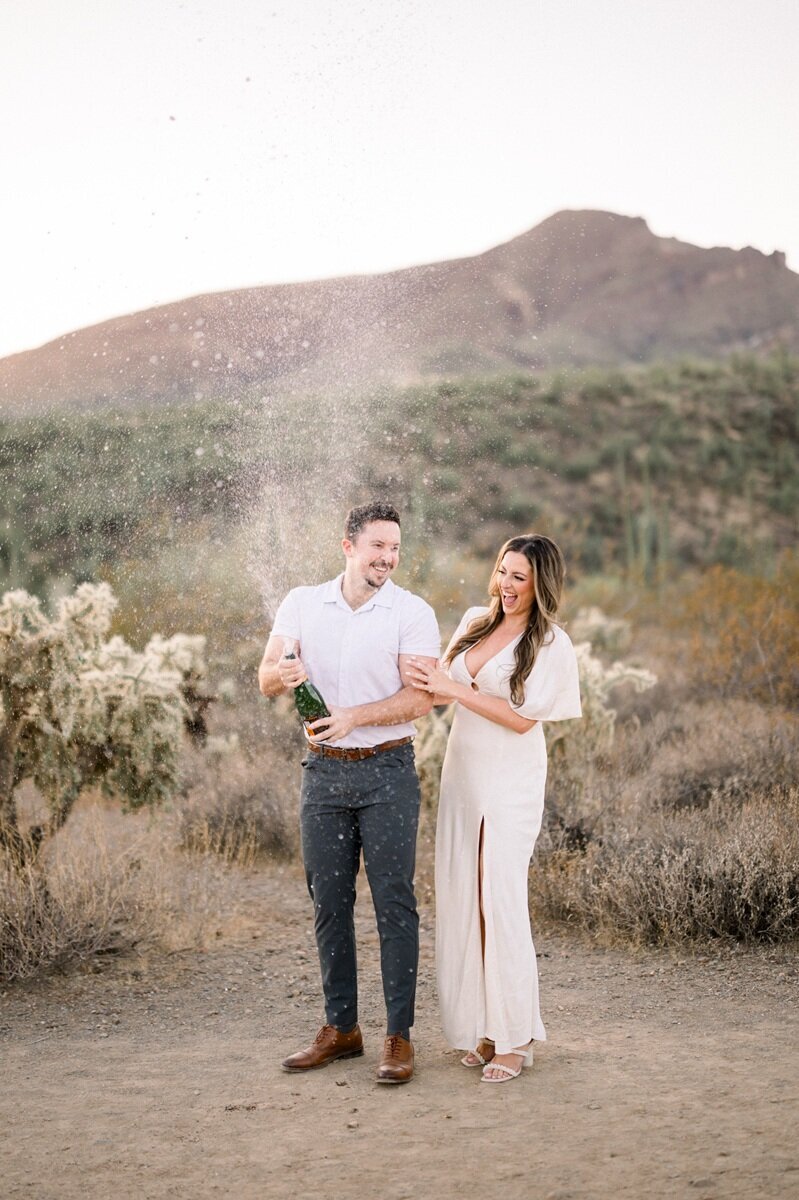 A couple standing together in a desert landscape with mountains and cacti in the background, dressed in coordinating white and neutral tones.