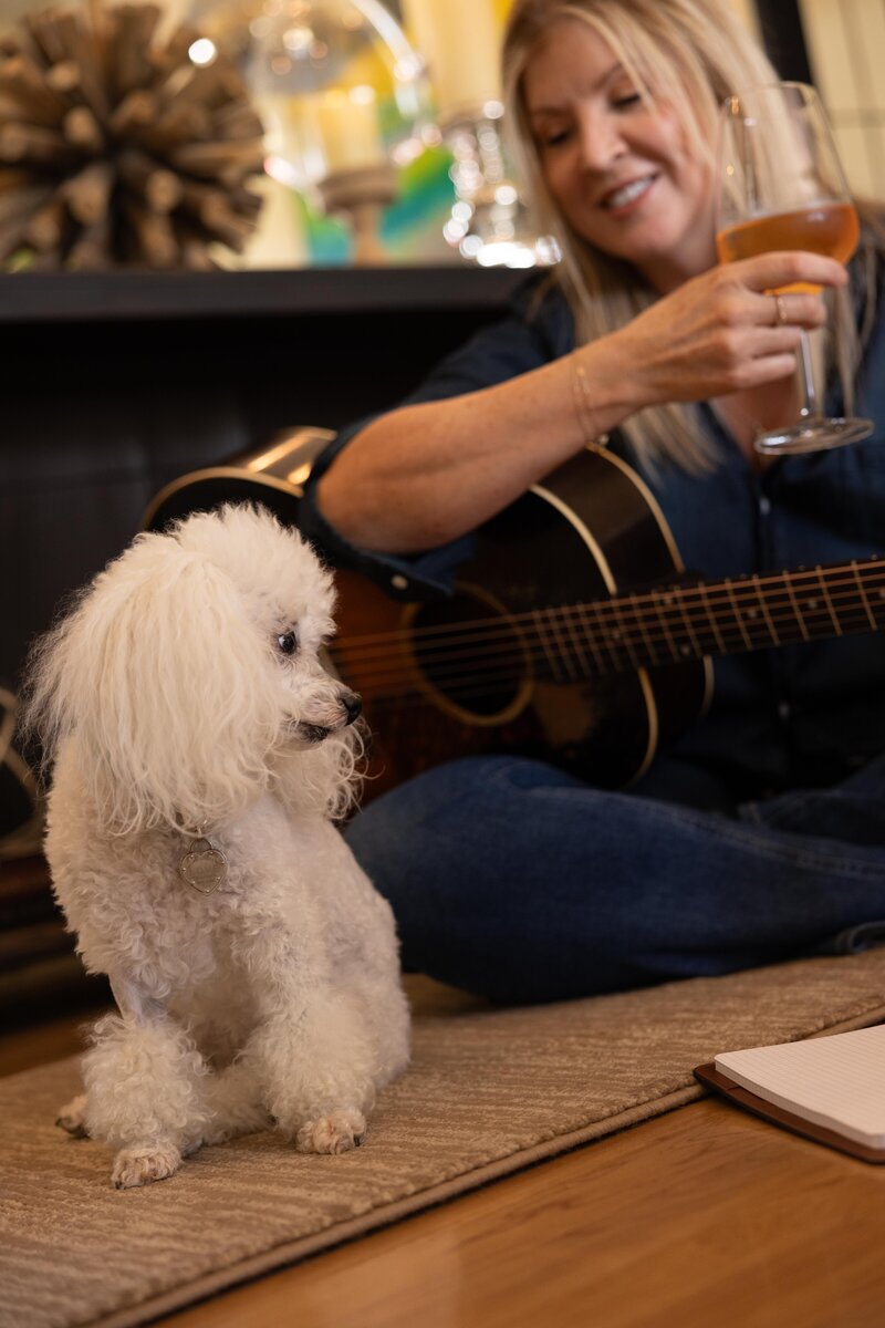 Amy sitting with acoustic guitar and poodle drinking a glass of wine.