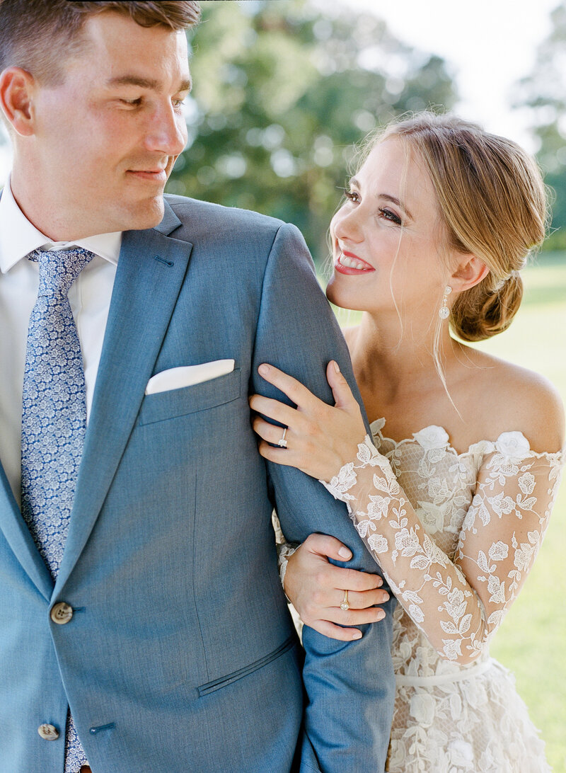 Bride and groom. The bride is wearing a floral lace off the shoulder dress, she is holding her grooms arm and smiling at him.
