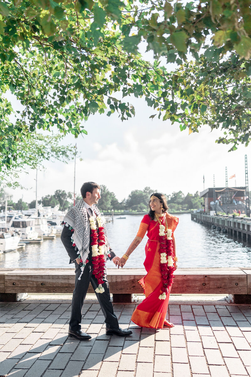 A couple in traditional attire, with floral garlands, hold hands and walk along a waterfront. They are smiling at each other, surrounded by trees and moored boats under a sunny sky.