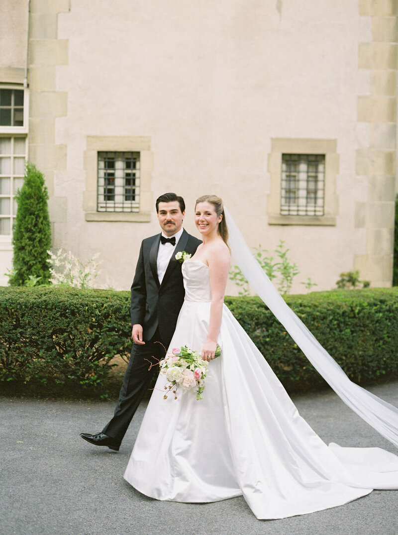 Glen Manor House | A bride in a white gown and a groom in a black suit walk happily on a path beside a hedge. The background features an elegant stone building.