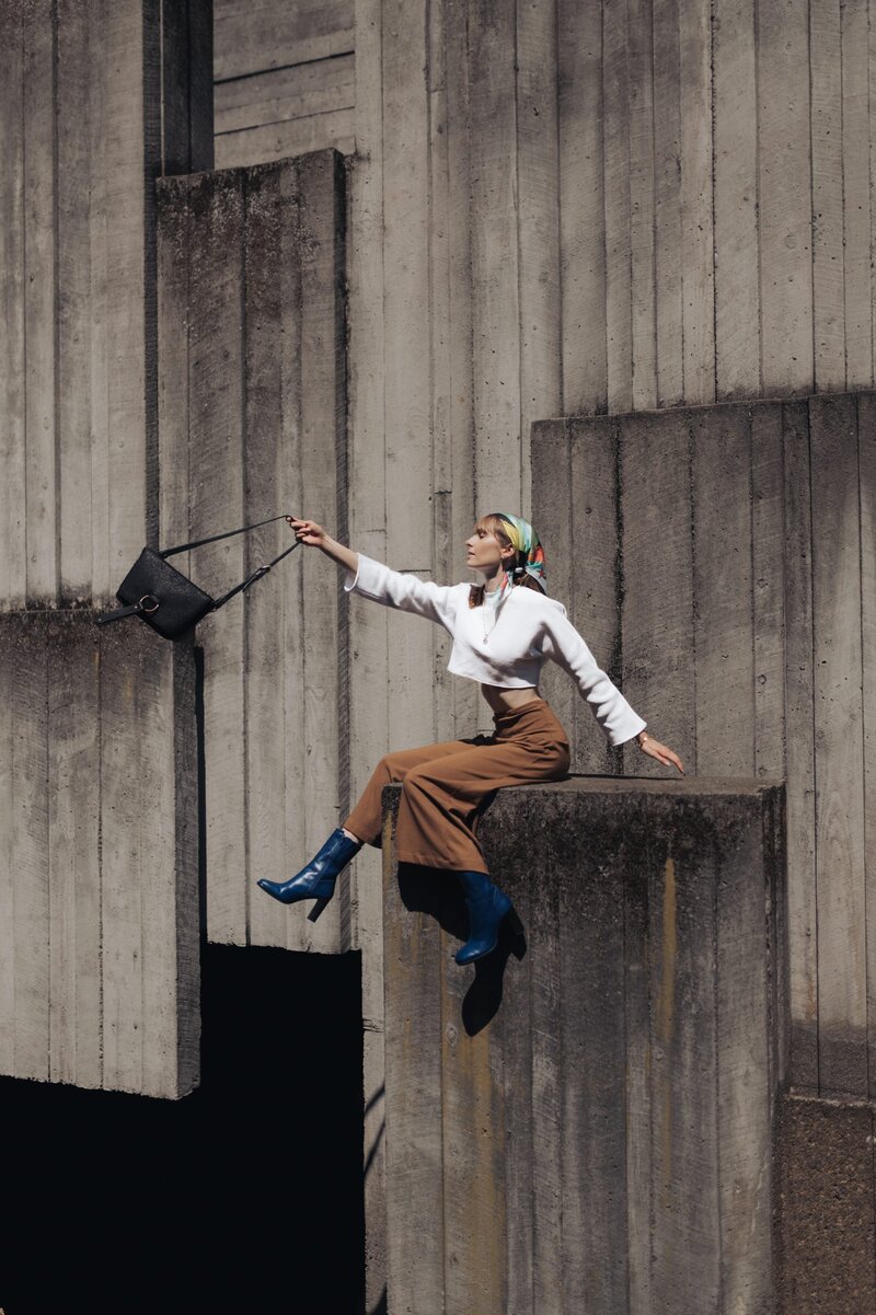 Woman sits on cement structure wearing a white shirt, brown pants, black heel boots and swings a black purse away from her.