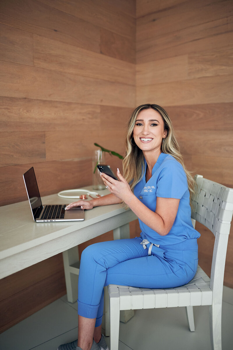 Mariya Kulyk holistic nurse coach dressed in blue scrubs smiling, working at her laptop.