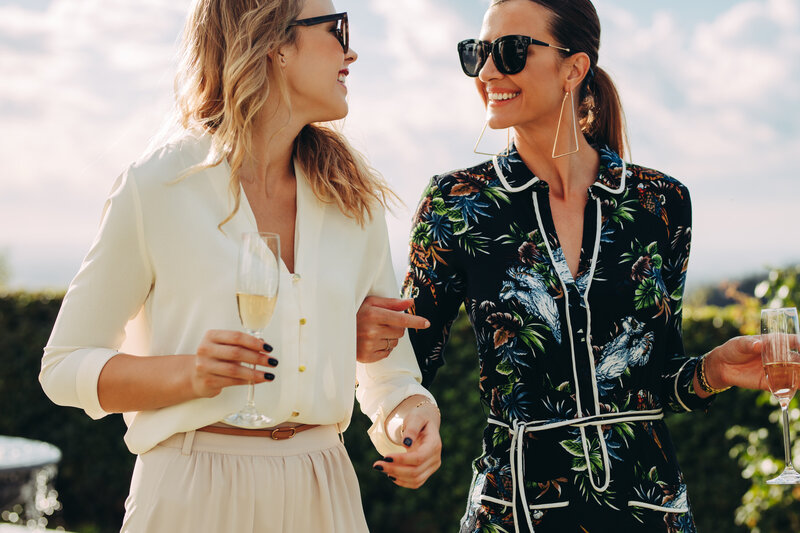 Two women smiling at each other while holding champagne glasses. One wears a white blouse and light skirt, the other a floral dress. Both wear sunglasses and appear to be outdoors in a sunny setting with greenery in the background.