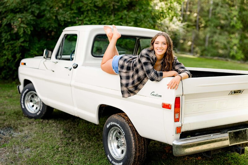 senior girl is posed on the back of a truck bed wearing  a flannel and jean shorts