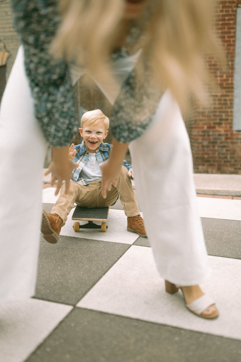 A mom reaches through her legs to pull her young son who is sitting on a skateboard and giggling.