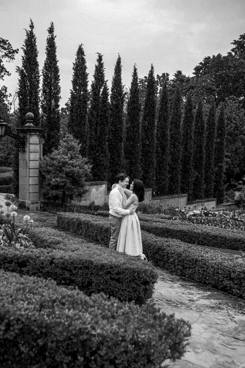 Engagement photo of couple embracing in the formal gardens at Philbrook Museum in Tulsa, Oklahoma surrounded by manicured hedges, cypress trees, and classic European architecture in black and white