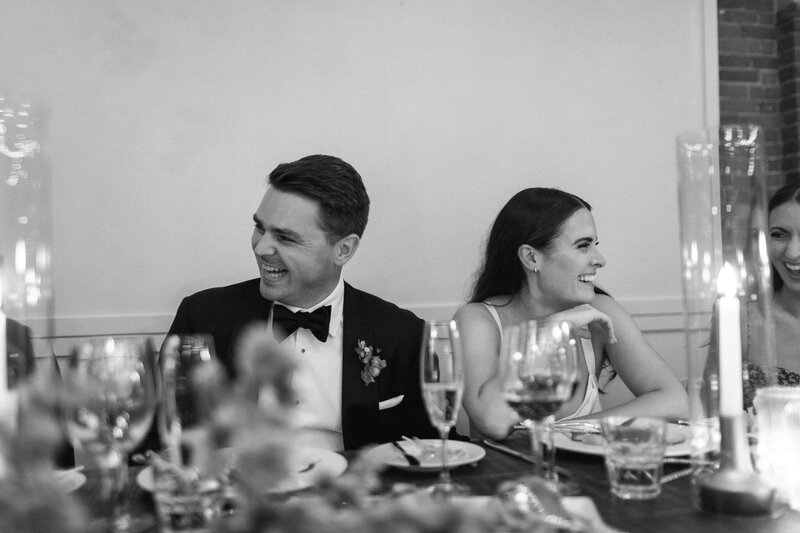 A group of people, elegantly dressed, share a laugh around a candlelit dining table at a Broadview Hotel wedding. Glasses clink amidst joyful chatter in this timeless black and white capture by Toronto wedding photographer, Jennifer van Son..