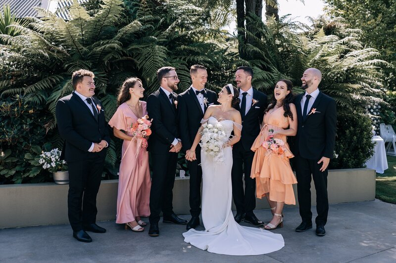 pink and peach mixed bridal party with black tuxedos and white florals stand in the garden at their christchurch wedding