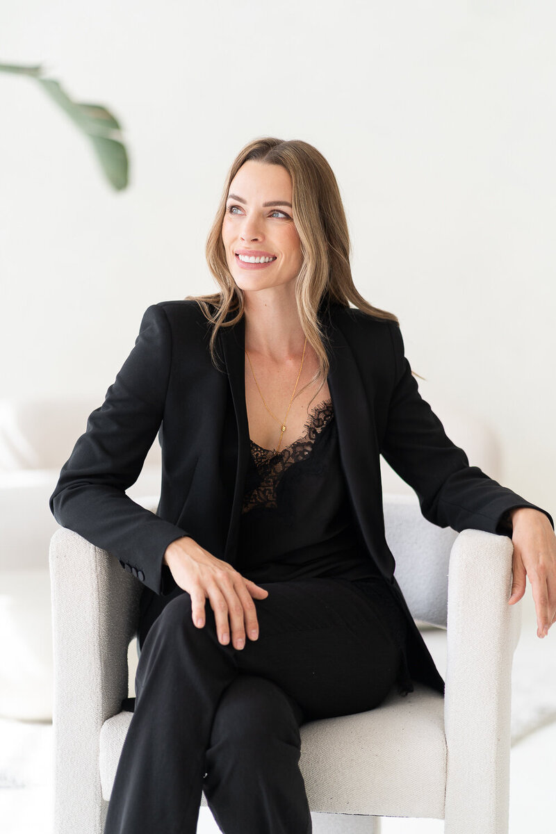 žena v černém kostýmku sedí v bílém křesle, usmívá se a dívá se doleva