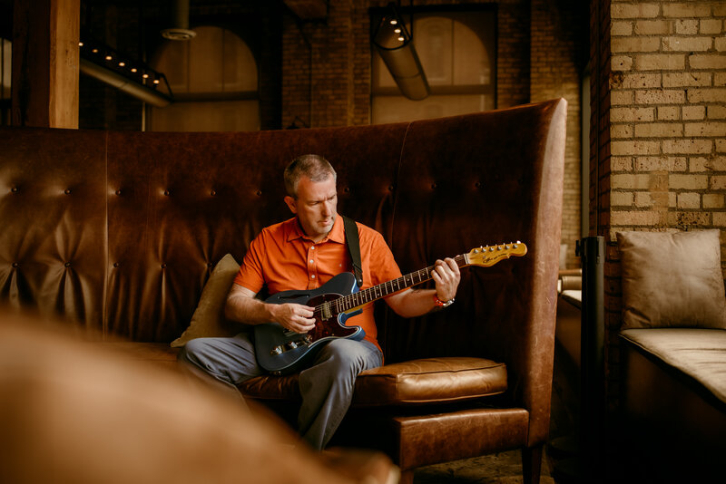 Man smiling during an online dating photo session in a modern indoor setting in Minneapolis playing his guitar