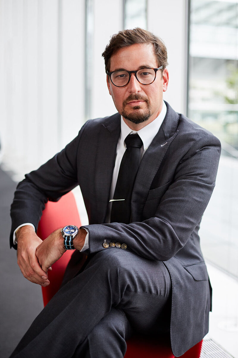 A headshot of a manager in a suit sitting on a red arm chair in a modern lobby