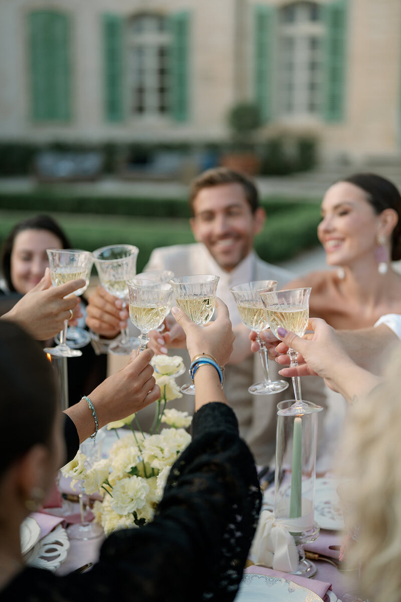 Bride, groom, and friends raising champagne glasses at a reception table, with the glasses in focus and the celebration blurred behind them.