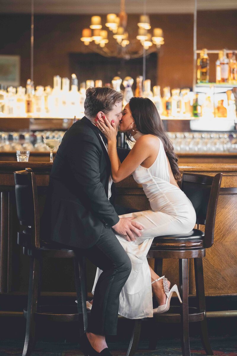 A bride and groom facing each other, kissing, at a bar