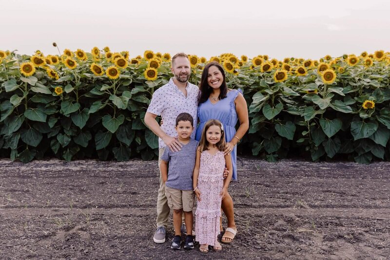 A family of four smiles in front of a sunflower field during golden hour.