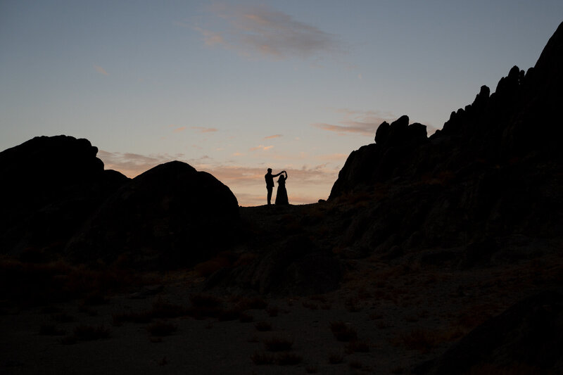 A couple dances on a mountain silhouetted against a darkening sky