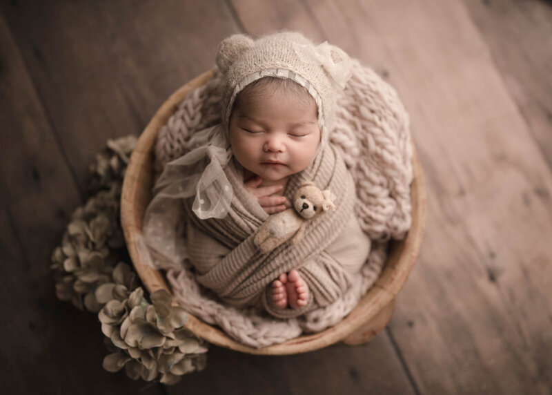 Aerial image. Newborn baby girl is posed for her Lake Elsinore Newborn Photoshoot. Baby girl is wrapped in taupe stretch fabric with her toes and fingers peeking out. She is wearing a taupe bonnet with bear ears and an organza bow under her chin draping over her side. Atop of her there is a tiny felt bear. Baby is sleeping peacefully. Captured by best Lake Elsinore newborn photographer Bonny Lynn Photography