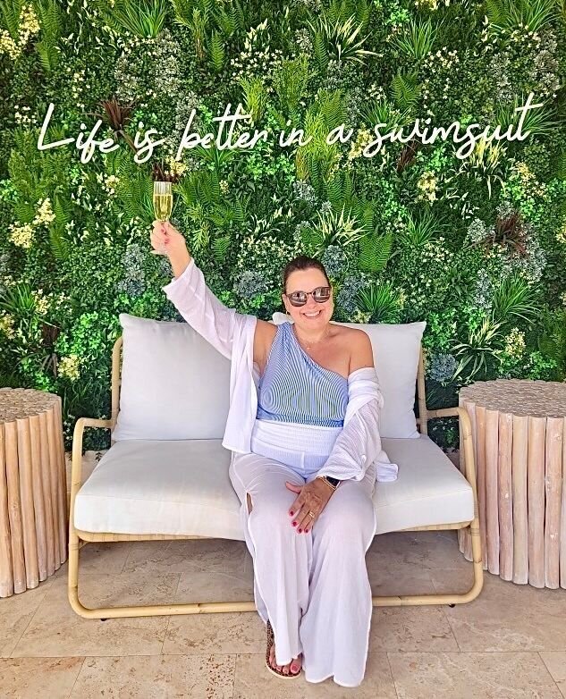 Female destination wedding planner wearing sunglasses and raising champagne glass in front of tropical greenery