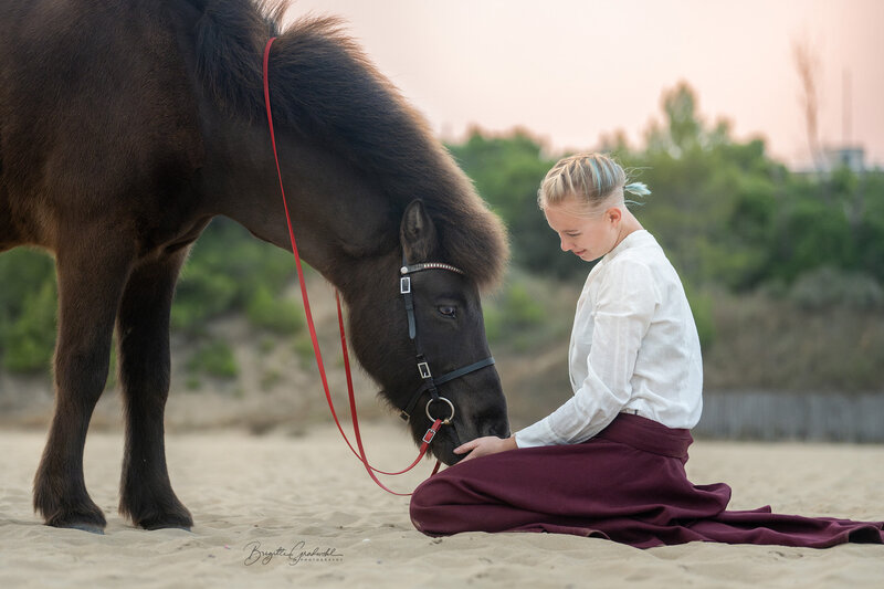 Pferdetrainerin und Pferdemassagetherapeutin in Salzburg, Deutschland, sitzt in einem roten Rock und einem weißen Hemd neben ihrem Islandpferd am Strand
