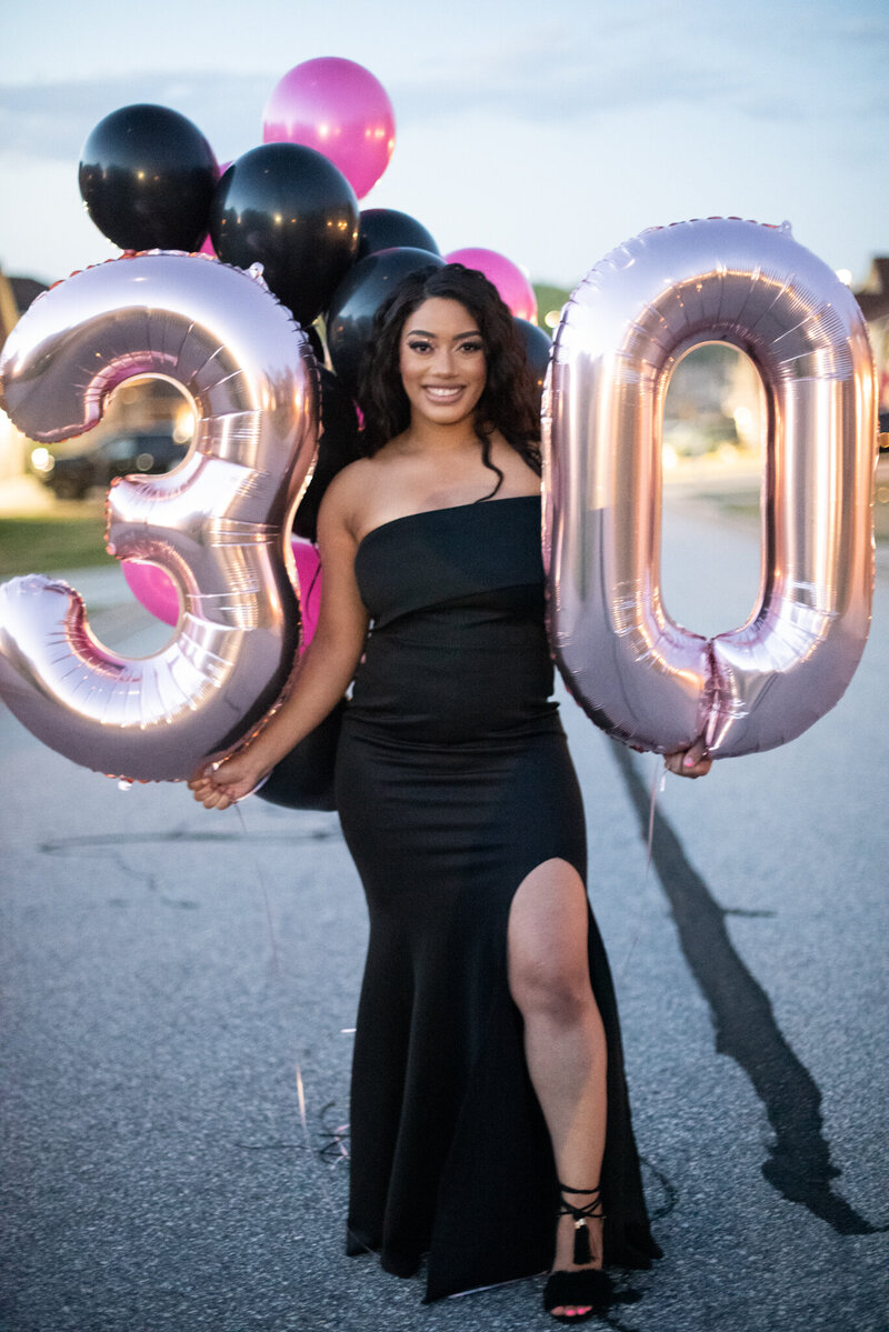 a woman in a black dress posing with two large balloons of the number 30 celebrating her thirtieth birthday. photographed by Millz Photography in Greenville, SC