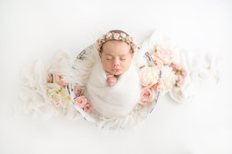 Newborn baby girl curled up and swaddled in a white antique bucket surrounded by pink and cream flowers