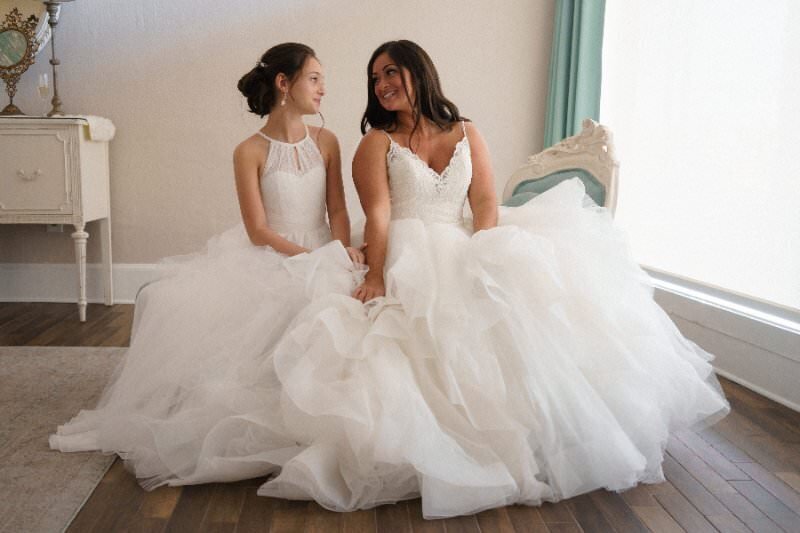 Two brides in elegant white wedding dresses sitting together on a bed in an ornate room with white vintage furniture and mint green accents at the White Room venue in historic downtown St. Augustine