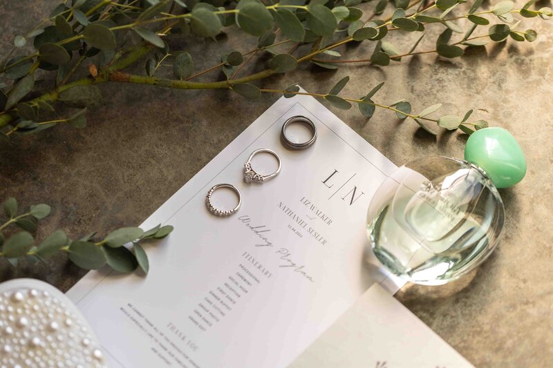 Wedding rings laying on top of wedding invitation suite with perfume bottle and wedding shoes laying nearby