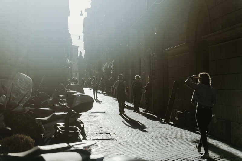 A sunlit italian city street scene with a woman taking a photo on the left, facing a row of parked motorcycles. Several people walk along the cobblestone street, bathed in the warm glow of the sunlight that filters through the narrow urban corridor. The architecture features classic stone buildings, adding a timeless quality to this busy, everyday moment.