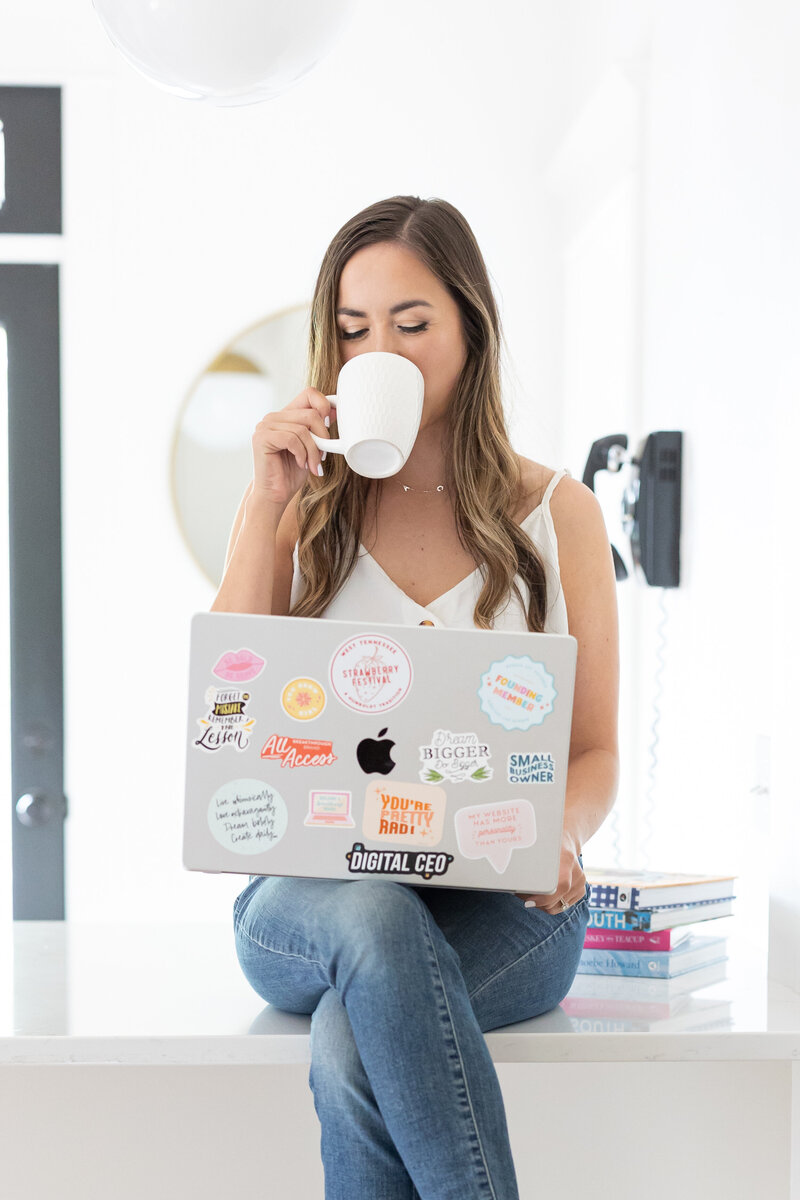 Woman during brand photoshoot sipping coffee and looking at laptop.