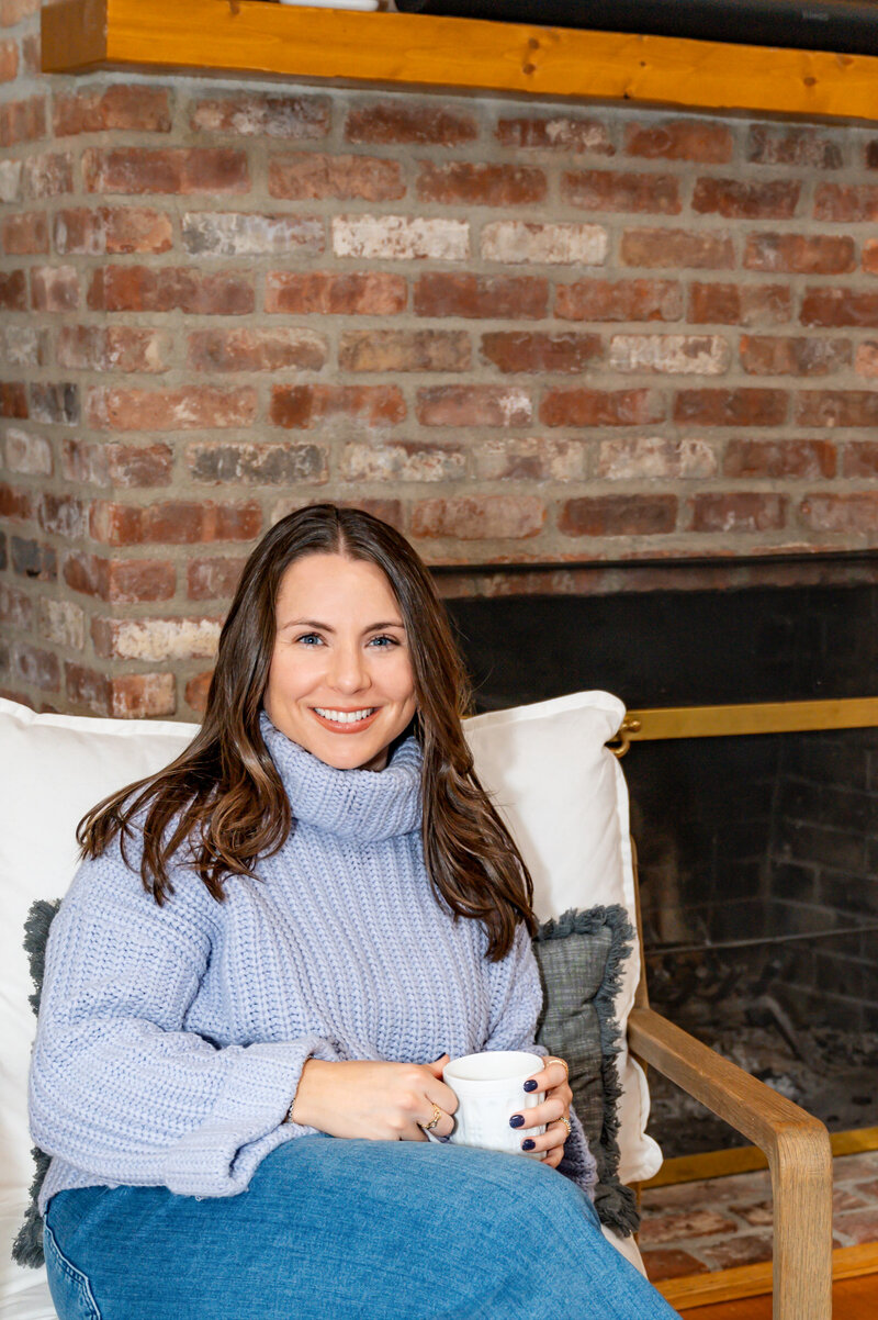 Brunette young woman in a blue sweater, sitting in a white chair, holding her cup of coffee, in front of a brick fireplace, posing for personal branding photos in Weston.