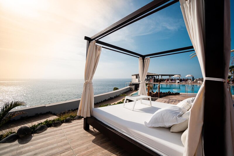 A luxurious outdoor cabana with white curtains and cushions overlooks a serene ocean. The sun sets in the distance, casting a warm glow. A nearby infinity pool reflects the vibrant blue sky, creating a tranquil and inviting atmosphere.