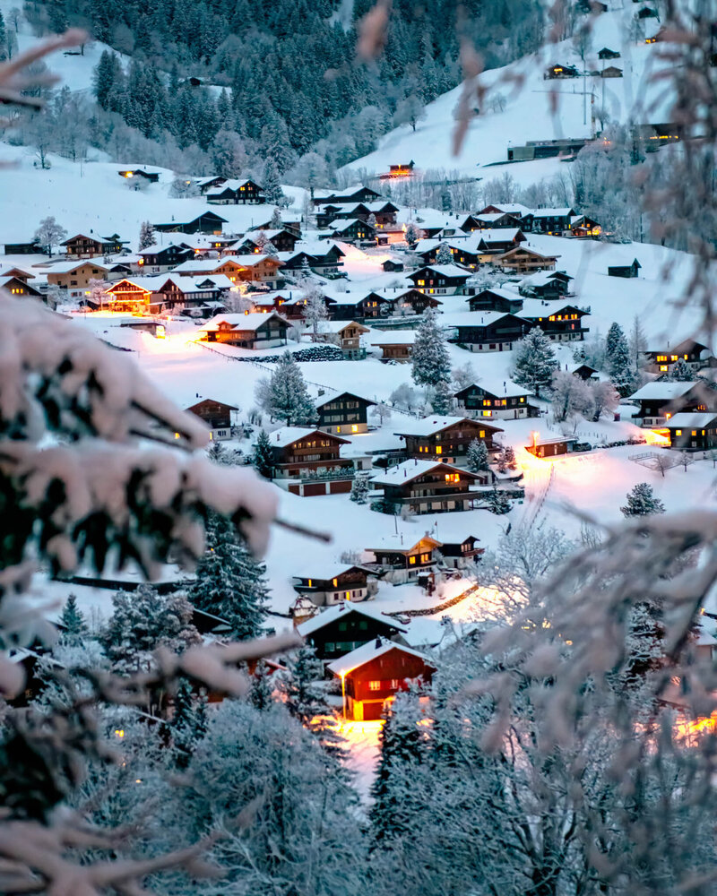 Snow-covered village nestled in a valley during winter. Warm yellow lights glow from wooden houses creating a cozy atmosphere. Snow-laden trees frame the scene in the foreground, with more forested hills visible in the background.