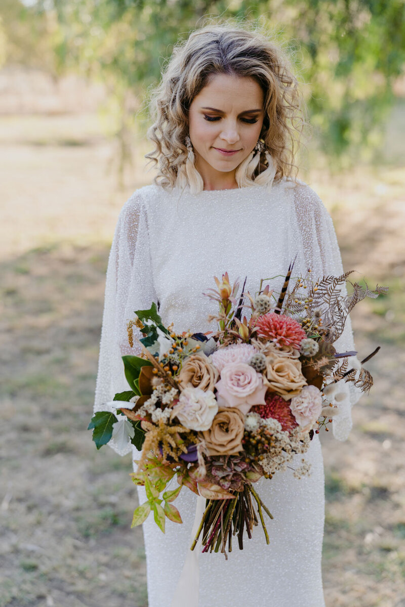 Bride in beaded dress with sleeves looking at her dried bouquet with mustard, cream and pink and purple flowers