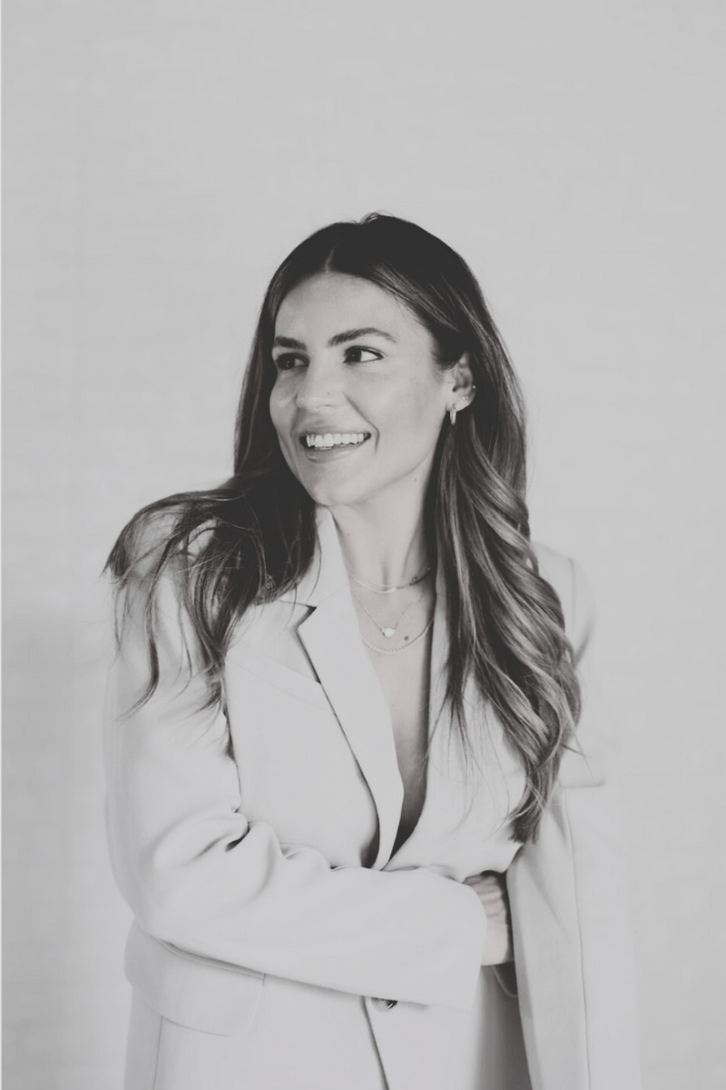 Natalie Bernacchi, a confident woman with long wavy brunette hair, wearing a light blazer and layered gold necklaces, smiling softly in front of a light textured background. 