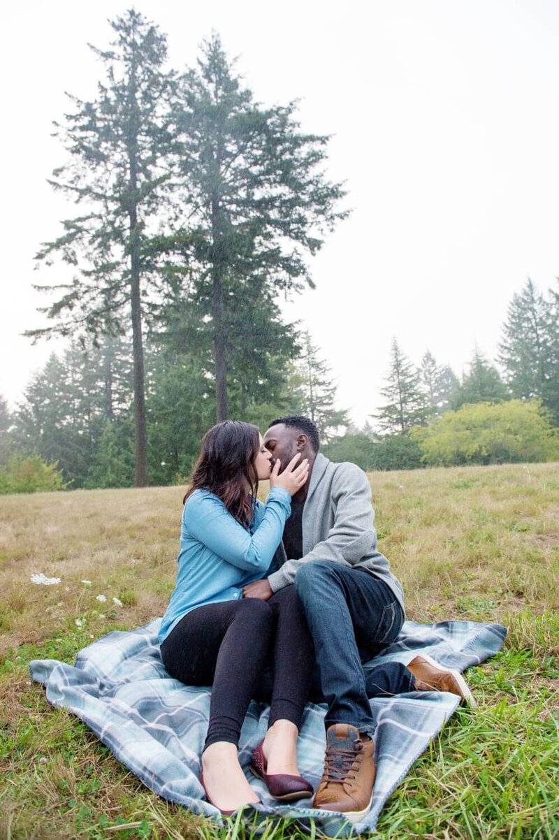 a man and woman sit in the grass in the rain and kiss at hoyt arboretum