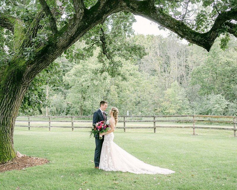 A newlywed couple stand together under a big beautiful oak tree after their Mayowood Stone Barn wedding in Rochester, Minnesota.