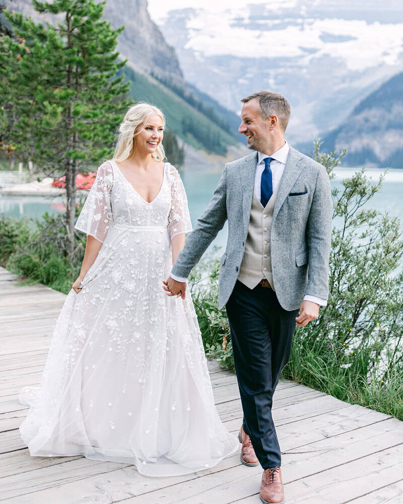 Newlyweds walk hand in hand along the scenic shoreline of Lake Louise, with the charming boathouse in the background, capturing the beauty of their special day.