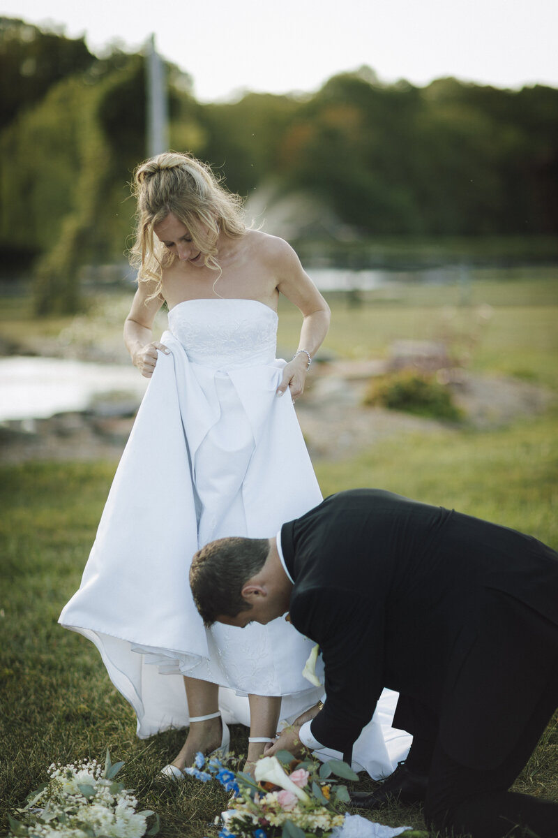 Groom kneeling to adjust bride’s dress beside a pond, a candid and caring wedding moment captured outdoors
