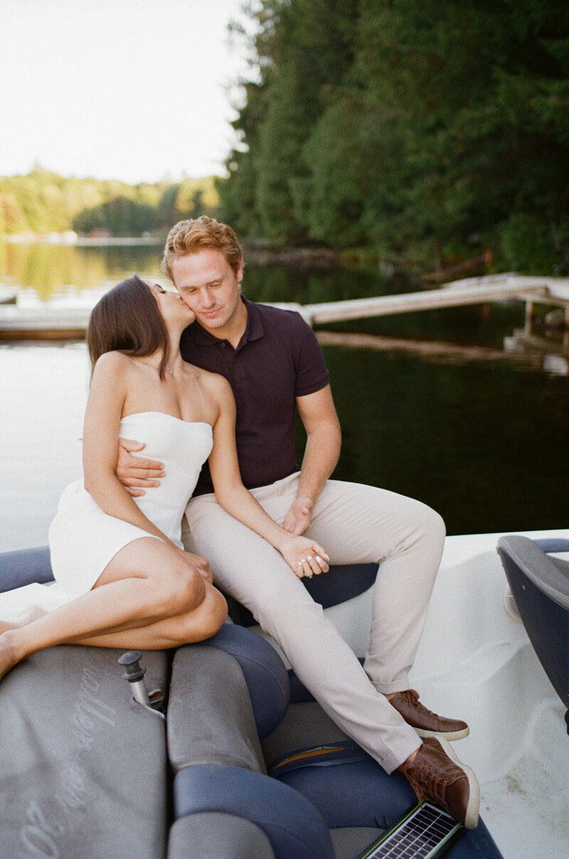 A couple sitting on a boat by a dock, with the woman in a white dress leaning towards the man in a dark shirt and light pants. Captured beautifully by engagement film photographer, Jennifer van Son, they are surrounded by a serene lake and lush trees.