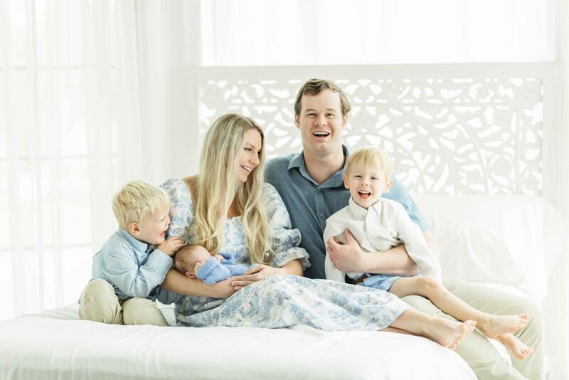 family of five sitting on a white bed in houston studio to hold their baby