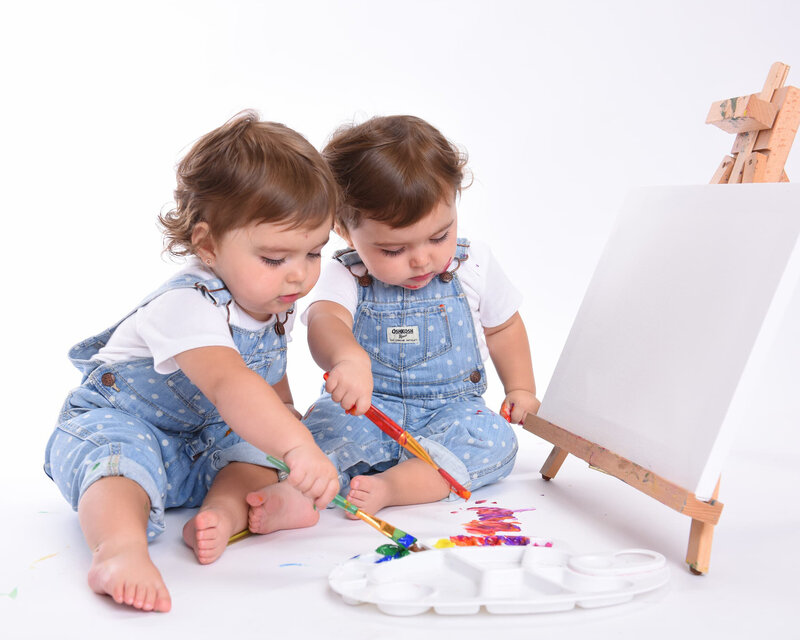 Twin toddlers in blue overalls painting on a canvas together