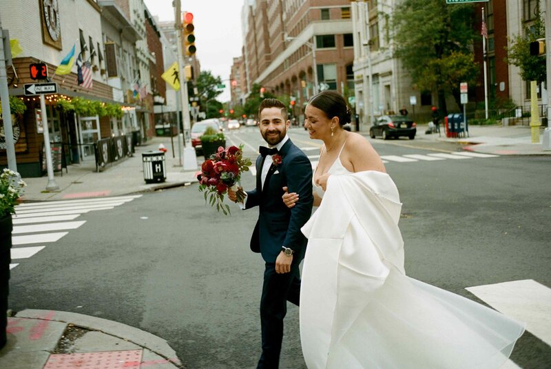 A stylish couple walks hand in hand through the streets of Hoboken, NJ, after their city-chic wedding, with the bride's modern gown flowing behind her.