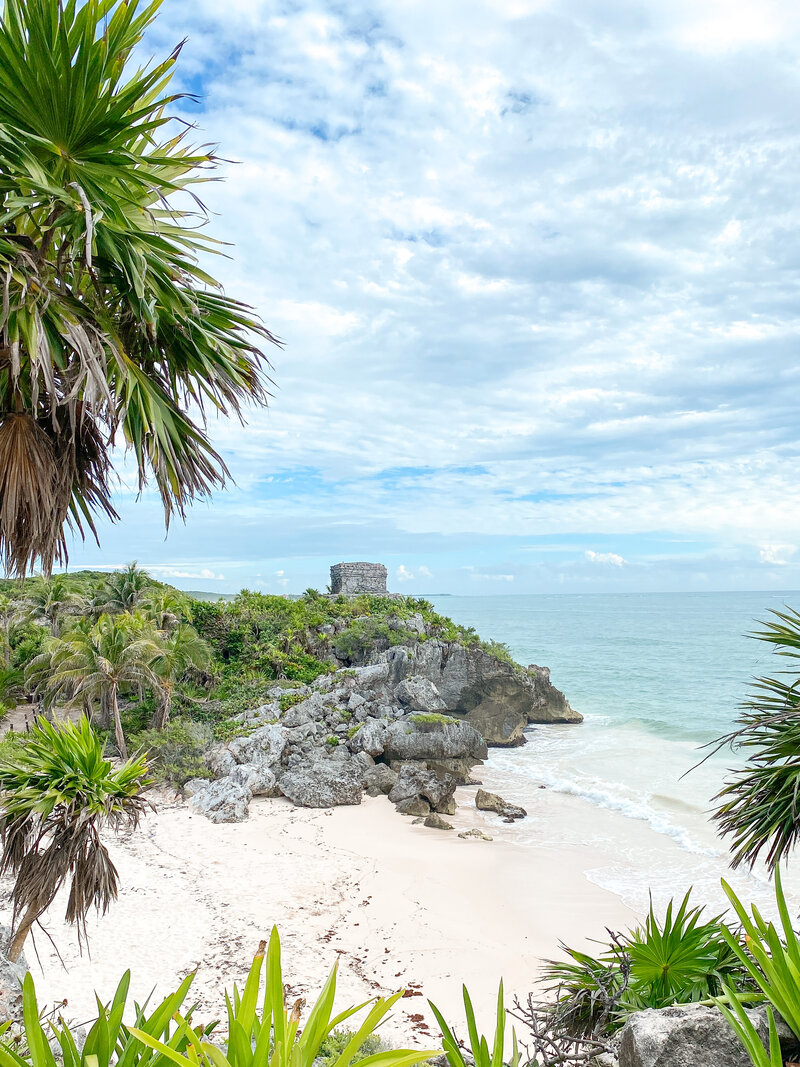 Scenic view of Tulum’s coastline, featuring lush greenery, rocky cliffs, and the historic Tulum ruins overlooking a calm, turquoise ocean under a bright, cloudy sky.