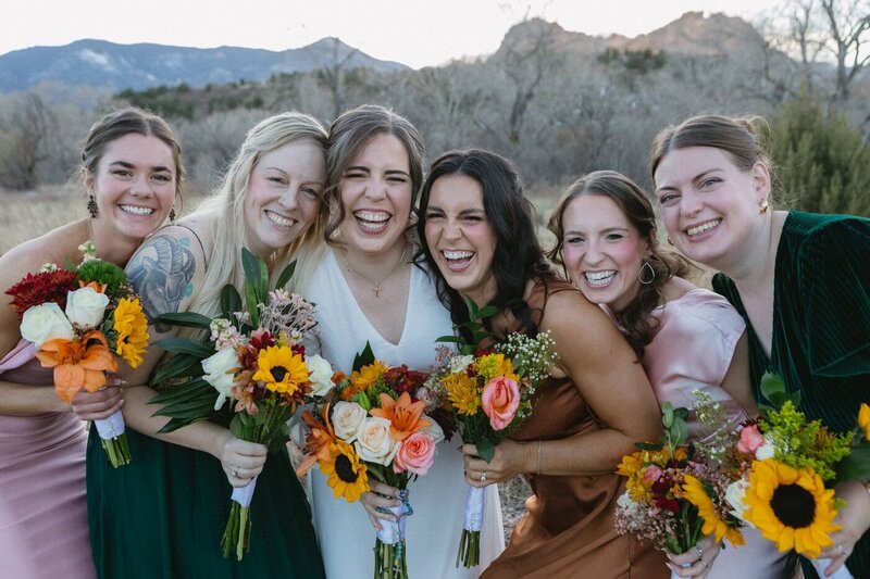 A bride and her bridesmaids stand huddled close together and laughing.