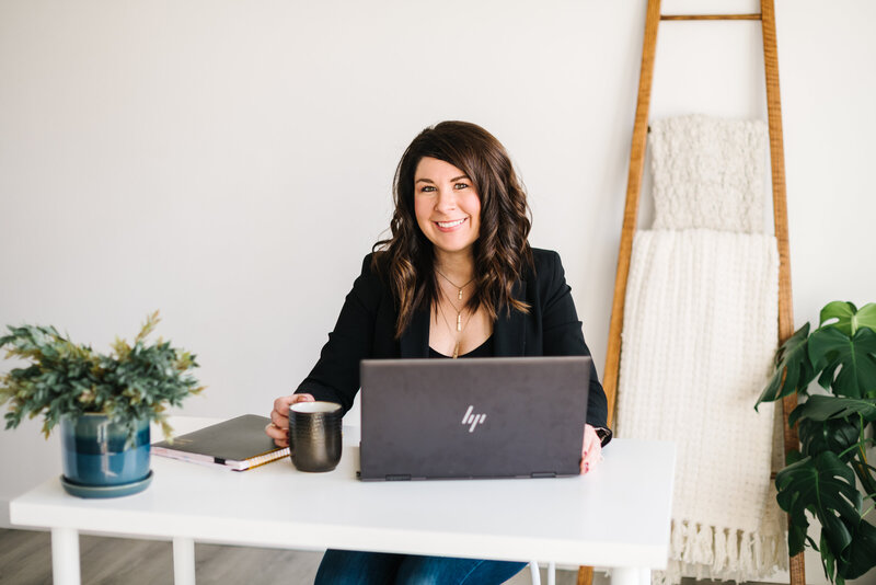 A smiling woman sits at her desk with her laptop and a coffee cup