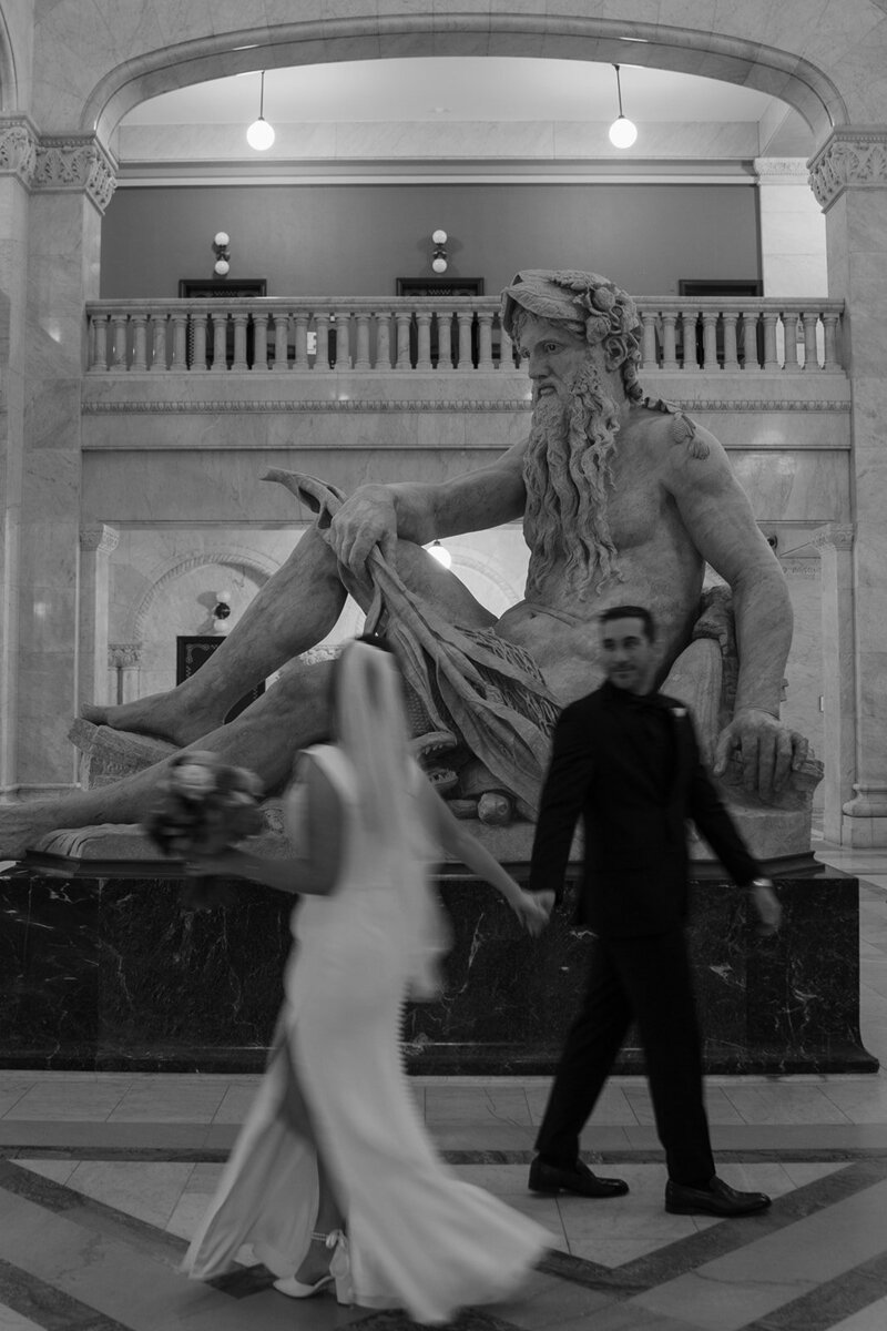 Bride and groom graze hands as they pass in front of statue of Greek god