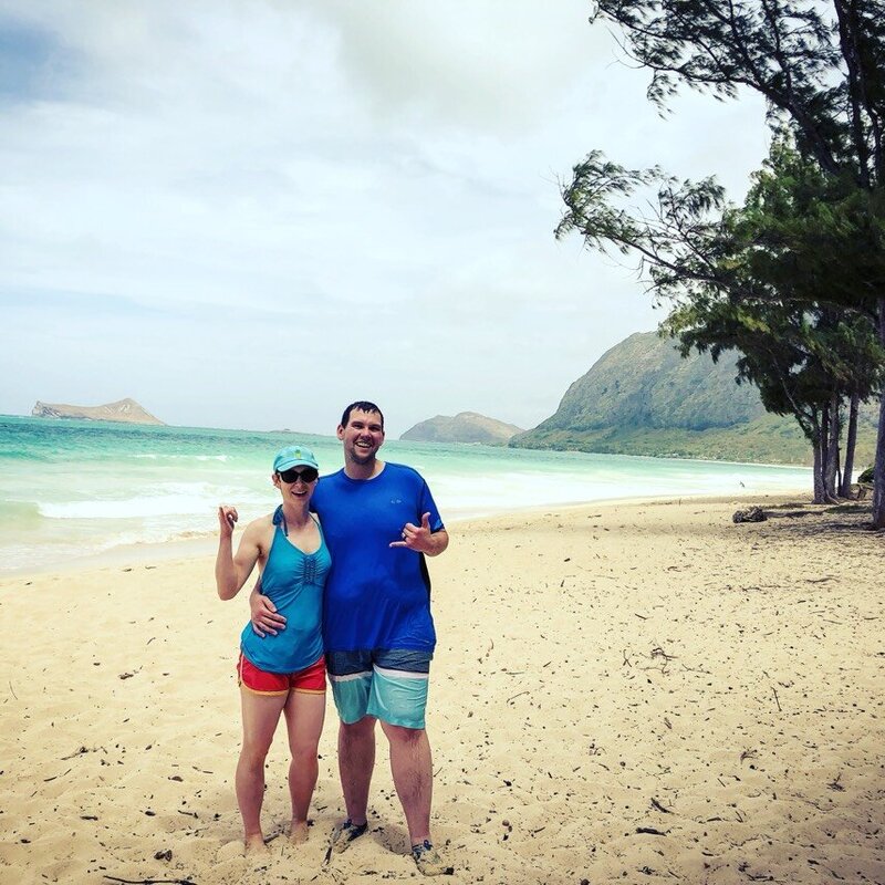 A couple stands on the beach of Waimanolo in Hawaii, smiling at the camera and giving shakabra