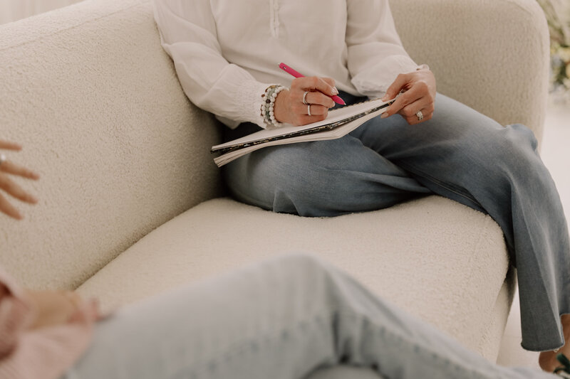 A woman sitting on the coach taking notes while wearing blue jeans and a white blouse